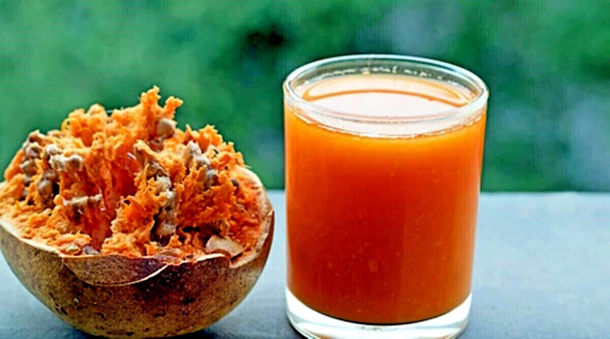 Glass of bael fruit juice next to a halved bael fruit with its juice extracted on a blurred green background