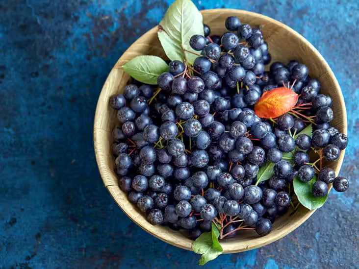 Wooden bowl filled with dark Chokeberries on a blue textured background. (Aronia melanocarpa)