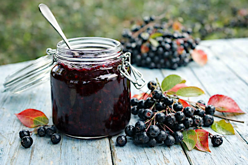 Jar of black Chokeberry jam with a spoon, surrounded by blackberries and leaves on a wooden surface. (Aronia melanocarpa)