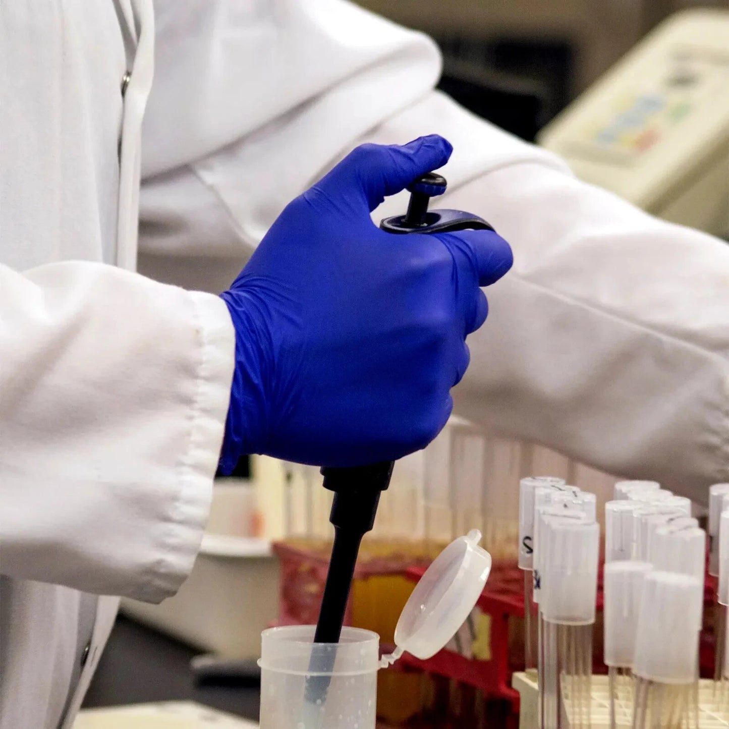 Person in a lab coat and indigo blue nitrile exam gloves using a pipette with test tubes in the background