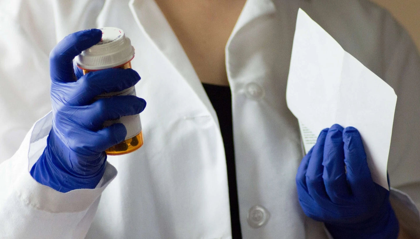 Person in a white coat holding a pill bottle and paper, wearing indigo blue nitrile exam gloves.