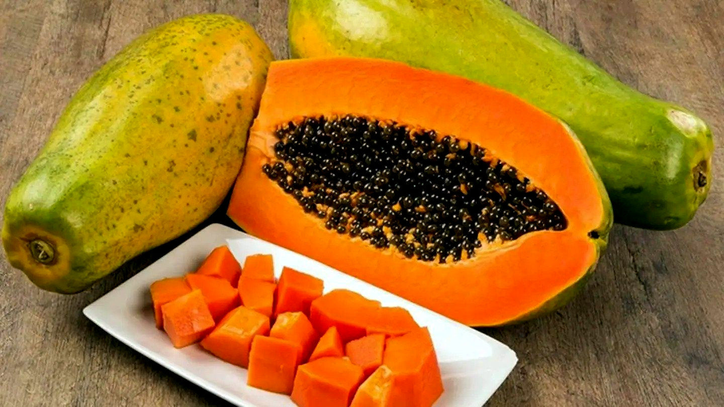 Papaya fruit with a cut open showing seeds, whole papayas on a wooden surface.