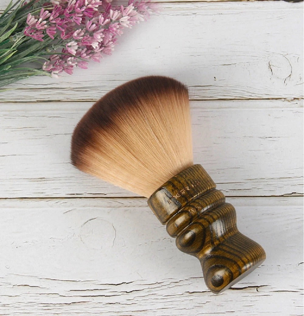 A shaving brush with a dark brown wooden handle and soft bristles that fade from dark brown to light beige, resting on a white wooden surface with pink flowers in the corner.