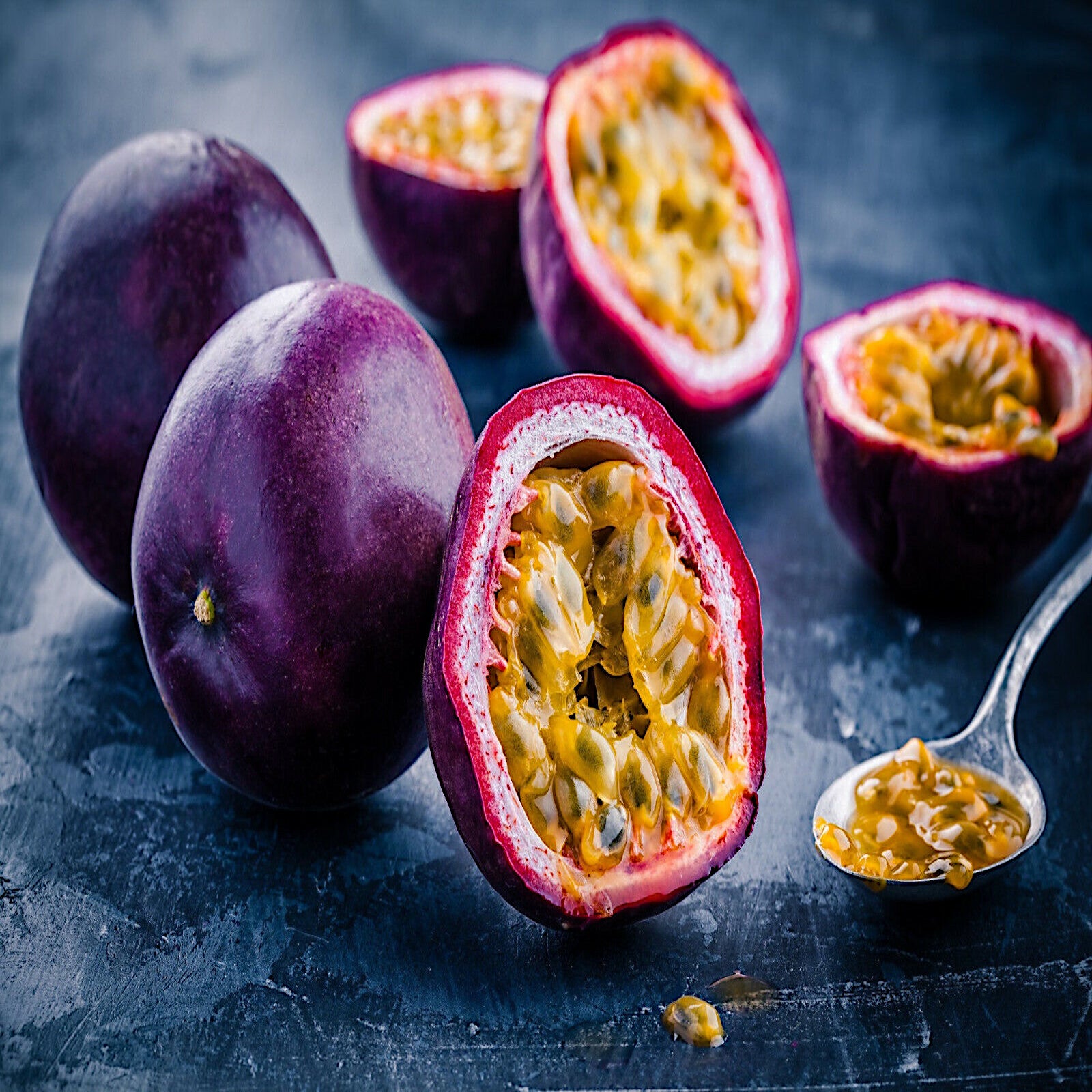 Purple passionfruit with one split in half with a spoonful of fruit lying on dark table.  Passiflora edulis
