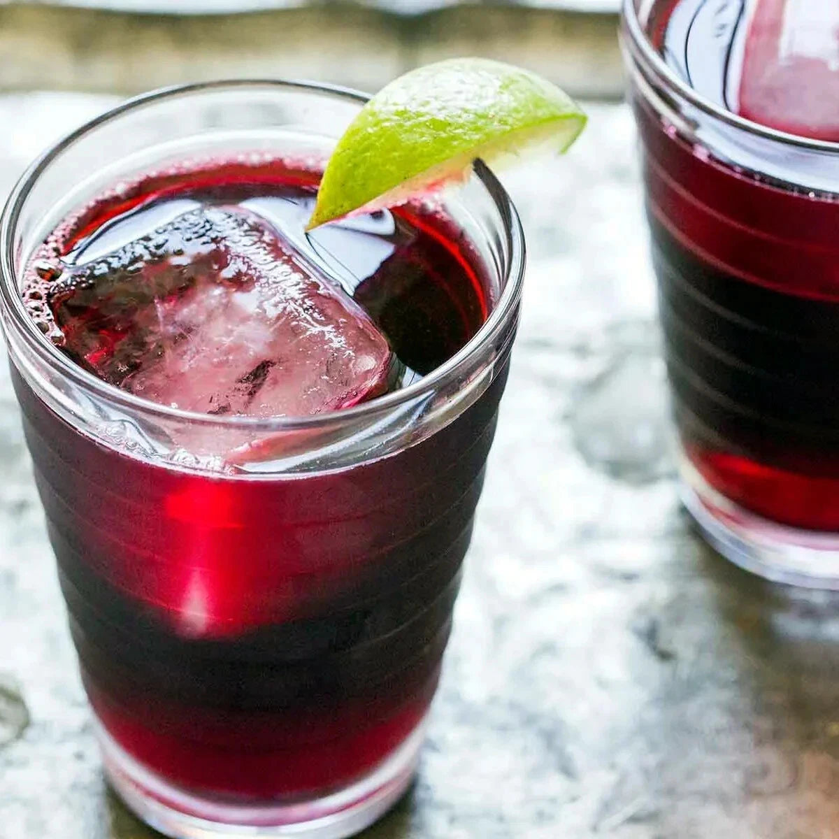 Glass of red Roselle juice drink with ice and lime wedge on a textured surface