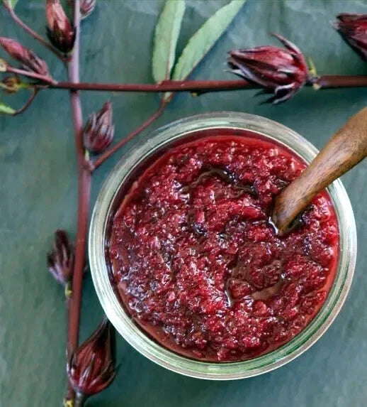 Jar of red Roselle hibiscus jam with a wooden spoon on a green background with hibiscus flowers.