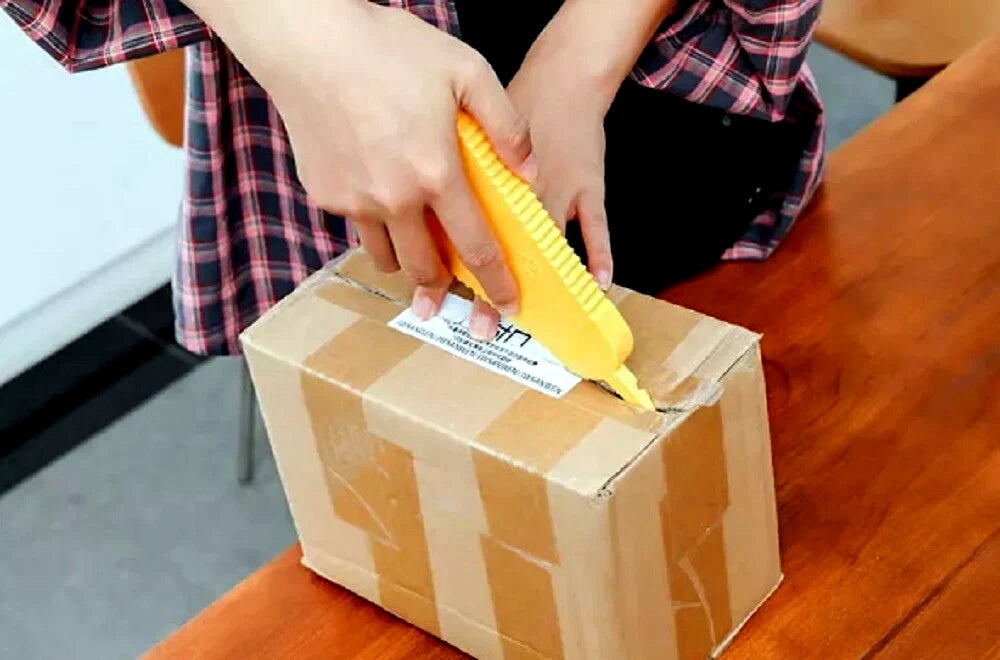 Person using a yellow multi function jar opener to cut seal on a cardboard box on a wooden table.