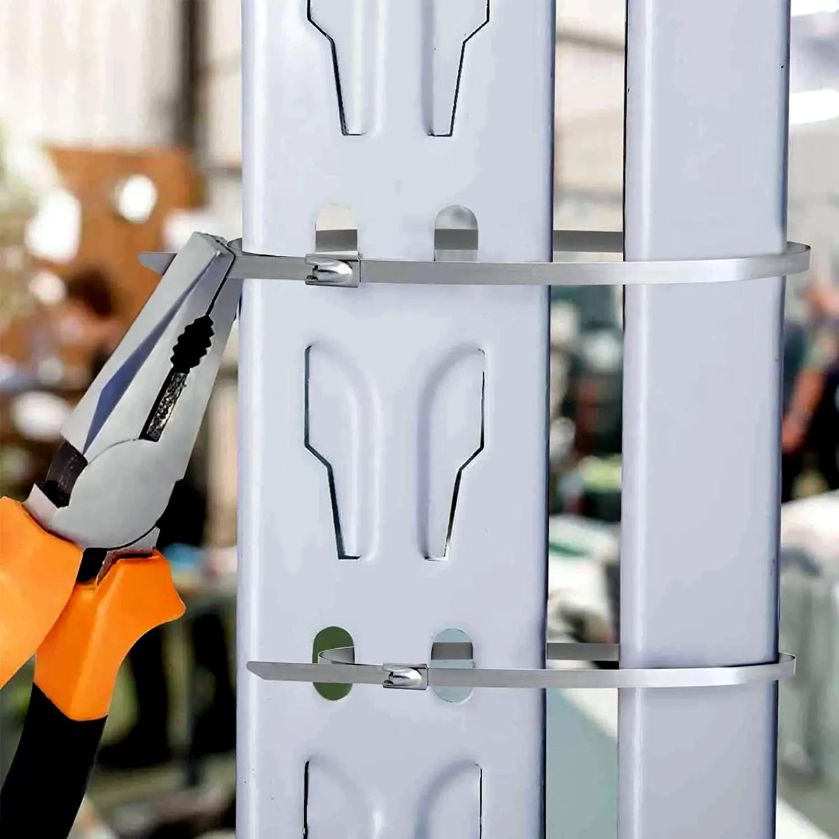 A close-up of silver metal cable ties securing white metal mounting brackets, with a pair of orange-handled pliers tightening one tie. The background is a softly blurred real-world setting.