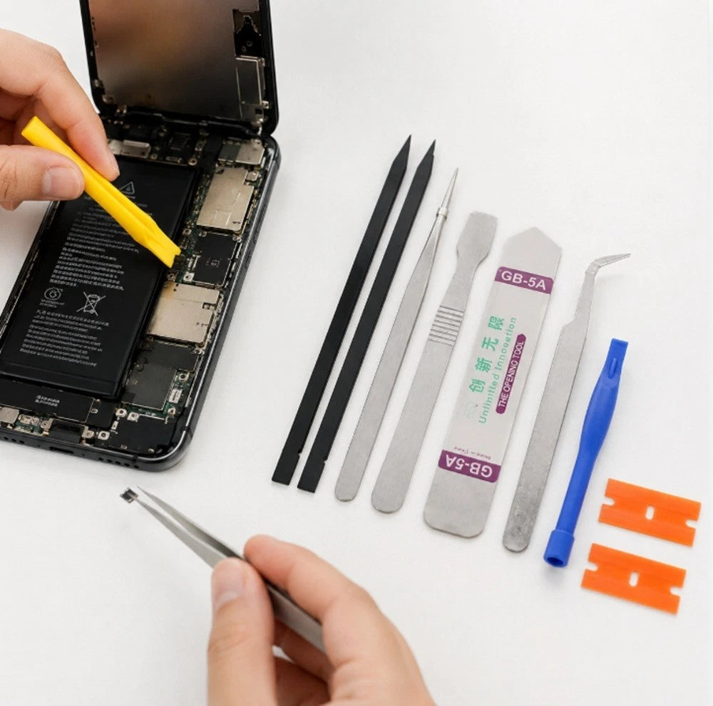 A close-up of a hand holding a smartphone disassembled to show the internal battery and circuitry, with a second hand using a yellow tool. Several small repair tools are arranged on a white surface: two black strips, two silver metal tweezers, a silver plastic spudger labeled GB-5A, a blue plastic hook tool, and two orange plastic spudgers.
