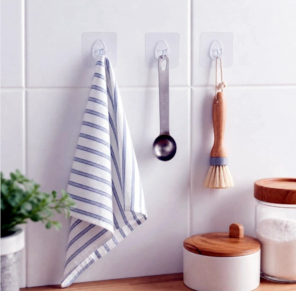Three clear adhesive wall hooks mounted on a white tiled kitchen wall, hanging a striped kitchen towel, a metal measuring cup, and a wooden-handled brush. A small plant and white wooden containers with a glass jar sit below on a wooden counter.